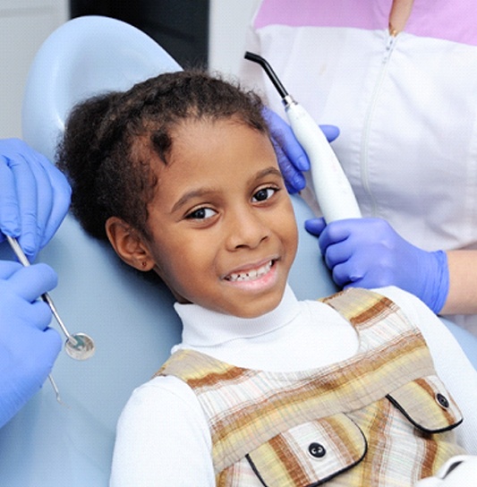 A little girl wearing a plaid jumper and white shirt smiles while a dentist and their assist prepare to check her smile