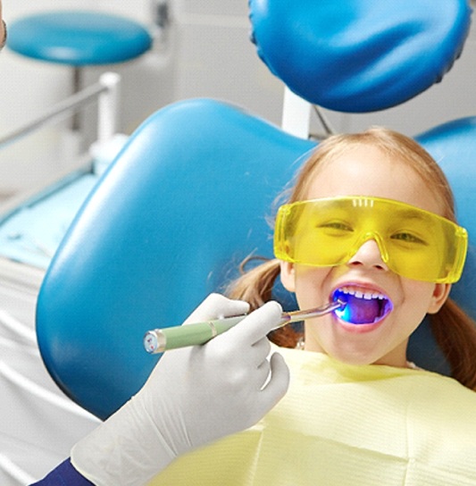 A dental hygienist uses a special light while applying a tooth-colored filling to a little girl’s smile