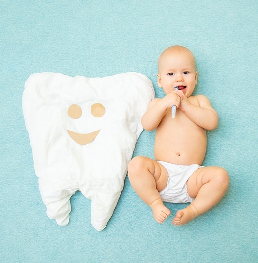 A baby wearing a pair of bloomers and brushing its teeth while lying next to a large tooth pillow