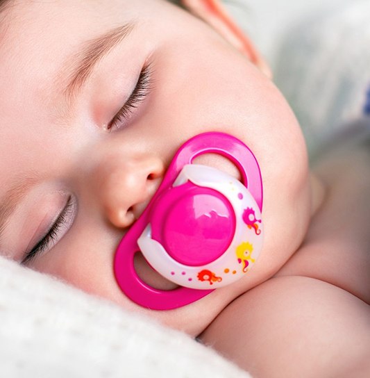 A baby asleep in bed with a pink pacifier in its mouth