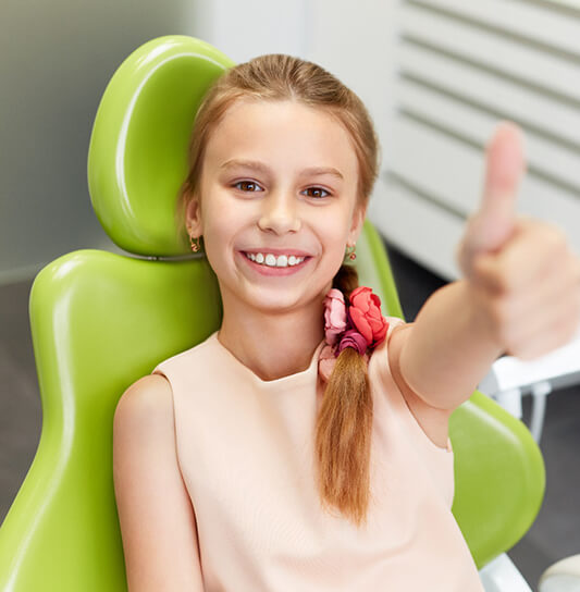 A young girl seated in the dentist’s chair and giving a thumbs up after receiving dental sealants in Cumming