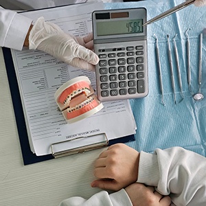 Bird’s eye view of gloved hand holding calculator next to model teeth with braces next to patient’s hands