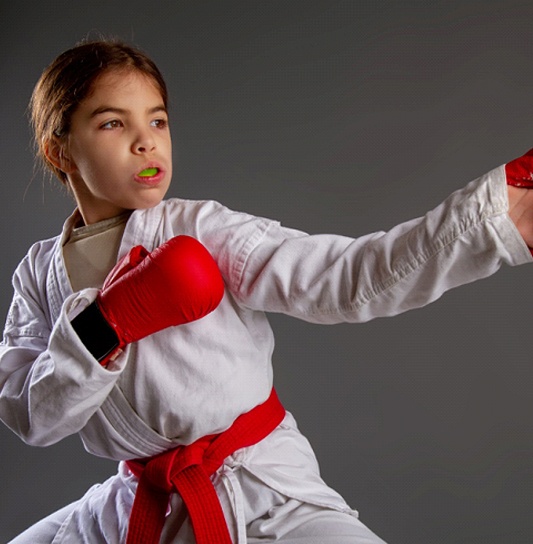 A young girl wearing a white robe, red belt, and red boxing gloves while protecting her mouth with a sportsguard