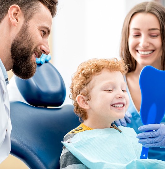 A little boy looking at his healthier smile in the mirror while the dentist and hygienist smile in the background