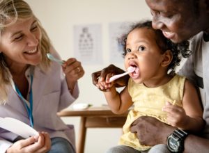 Toddler and her dad with a dentist learning how to brush her teeth together